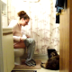 A girl sitting on a toilet and her dog battle for space in the bathroom.